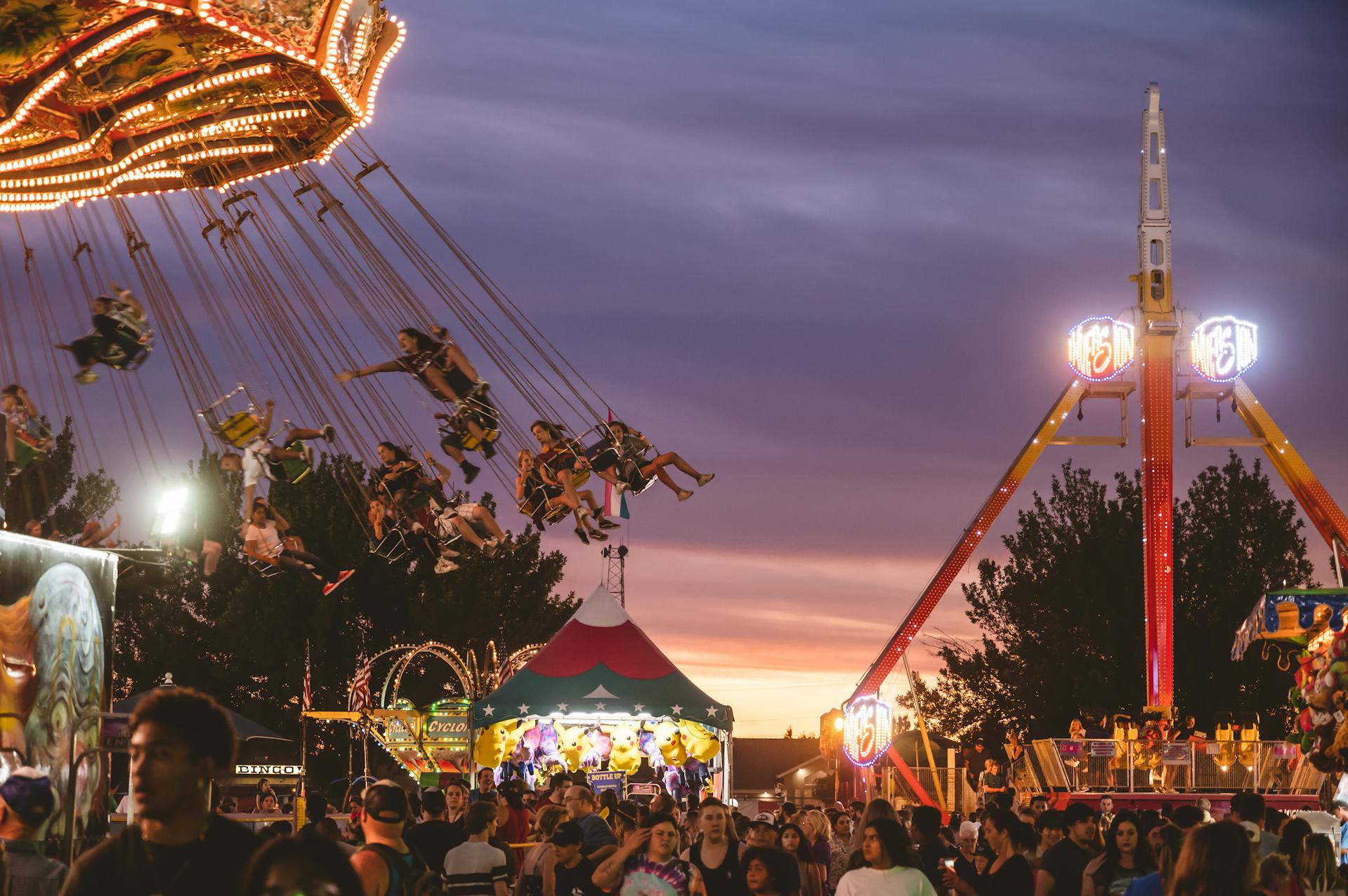 People enjoying a chair swing ride at an amusement park during dusk, with festive lights and a vibrant sunset in the background.