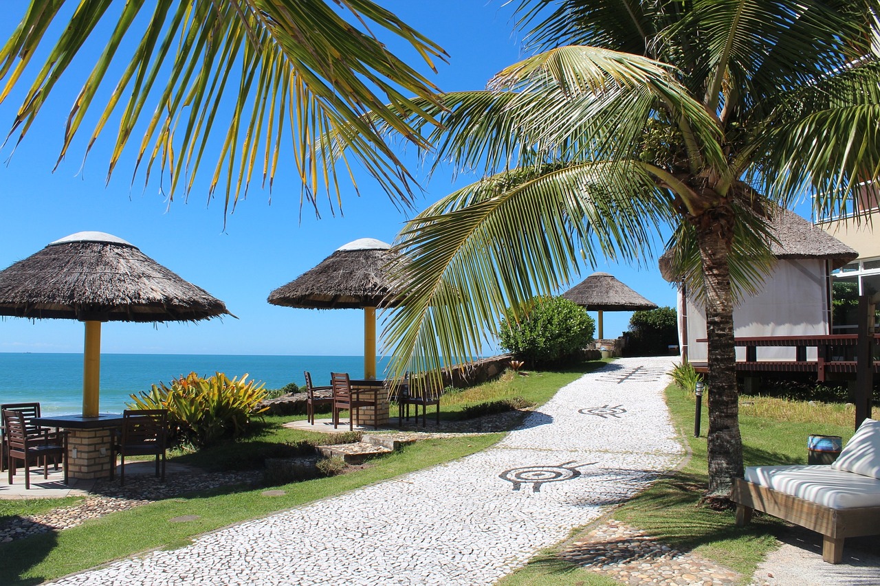 Relaxation area at a beachside resort with thatched umbrellas, palm trees, and pebbled paths leading to the serene blue ocean under a clear sky.