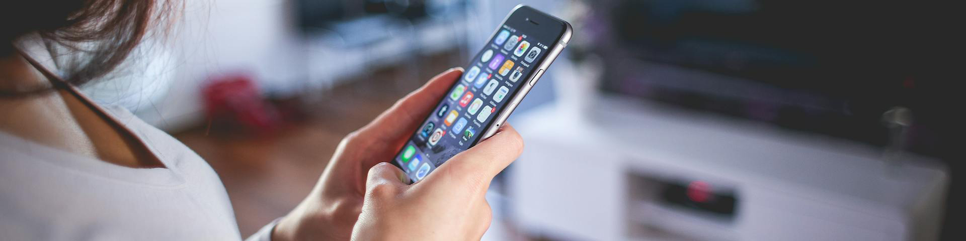 Woman in a white shirt browsing apps on a smartphone in a living room setting, focus on the smartphone screen.