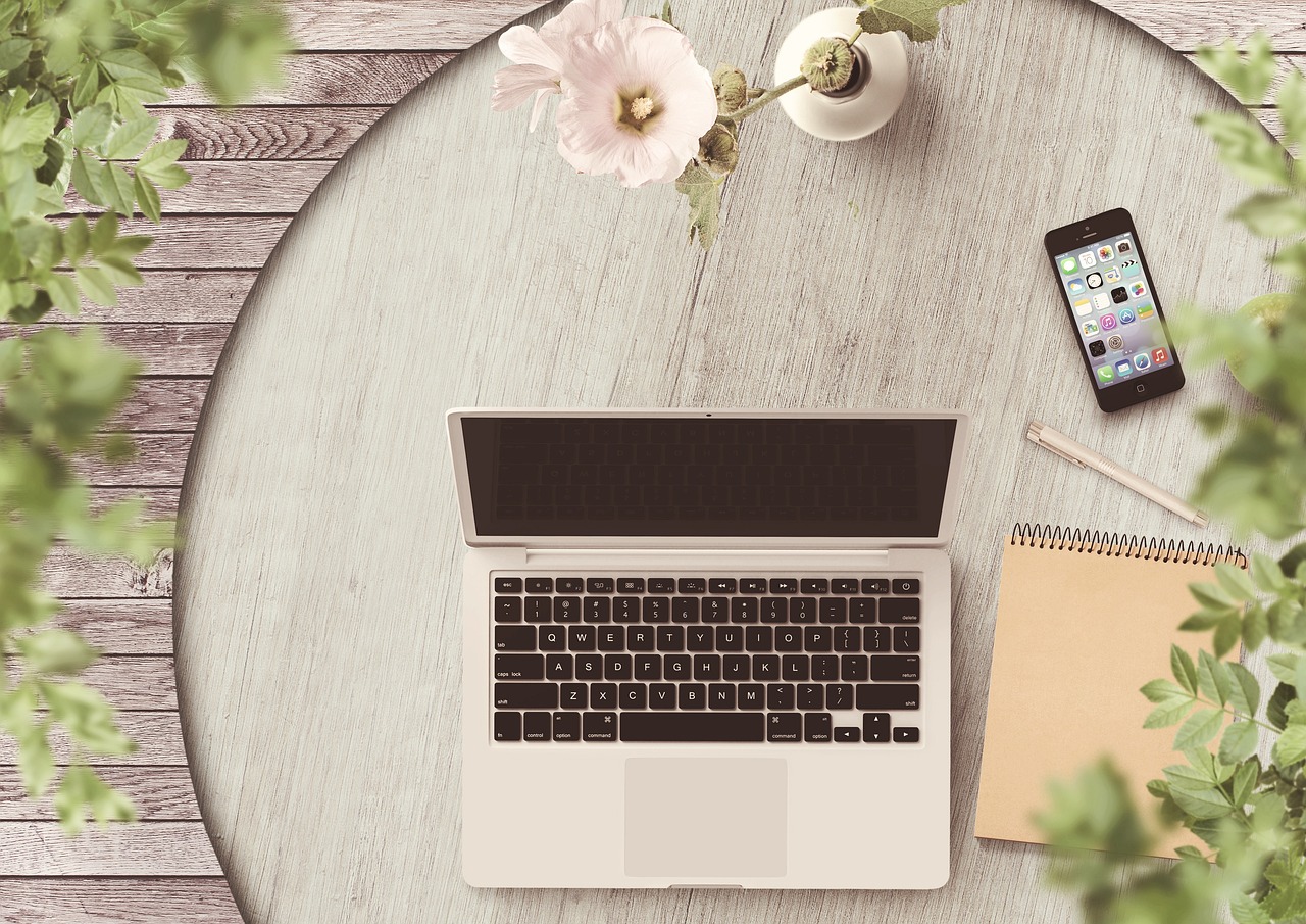 A top-down view of a home office setup with an open laptop on a round wooden desk, a vase with a blooming pale pink flower, a smartphone, and a notepad with a pencil on a grey wooden surface, surrounded by green foliage.