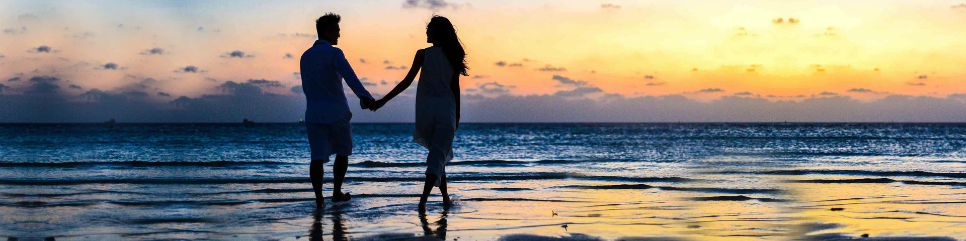 Silhouetted couple holding hands on a beach at sunset, with calm ocean waves and a colorful sky in the background.